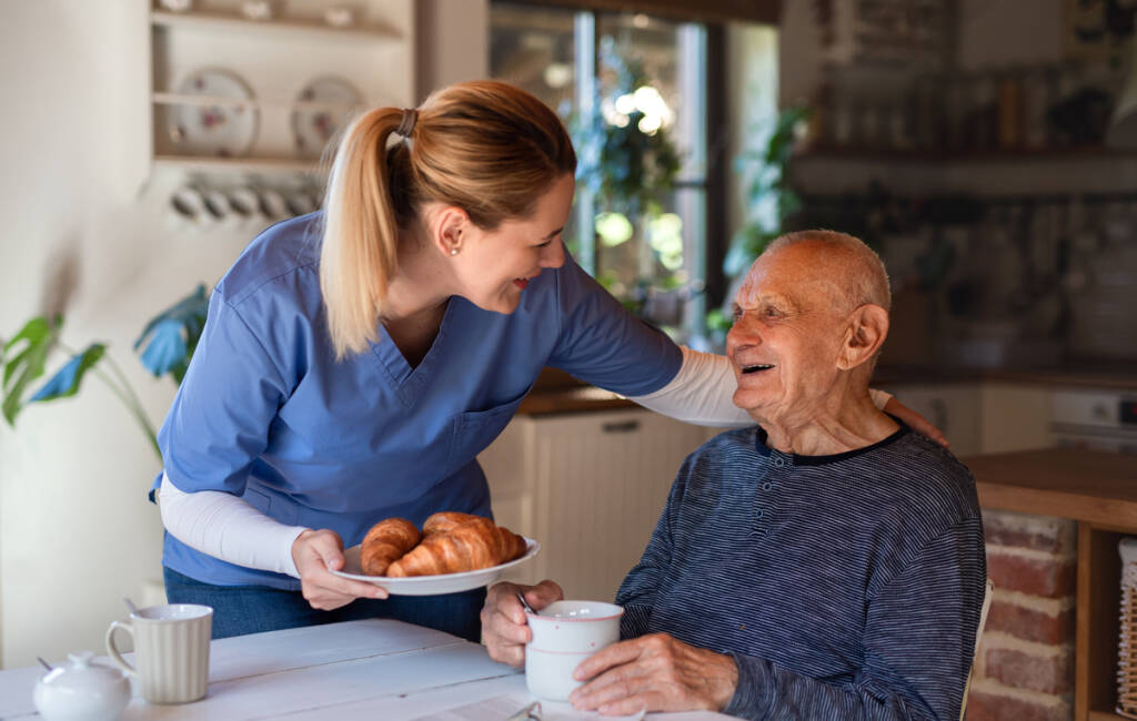 Happy young female caregiver or healthcare worker visiting senior man indoors at home, assisting with breakfast.